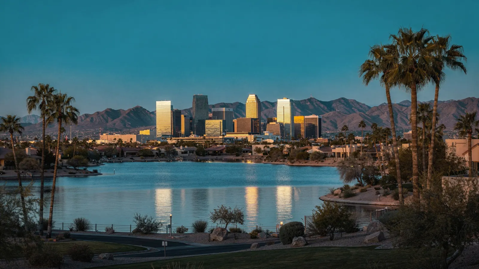 Palm-lined boulevard and a distant glass skyline across a landscaped desert lake at dusk